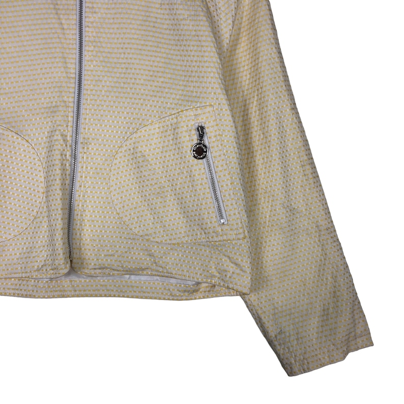 K&ouml;nnte beinhalten: Eine gelbe Jacke mit wei&szlig;em Muster, Rei&szlig;verschluss und einer Rei&szlig;verschlusstasche auf der linken Seite. Die Tasche hat ein kleines kreisf&ouml;rmiges Logo mit braunem Zentrum.