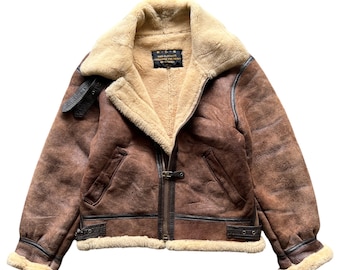 Puede incluir: Chaqueta bomber de cuero marrón con cuello y forro de borrego. La chaqueta presenta cierre de cremallera, detalles de hebilla y una etiqueta en el interior. La chaqueta tiene un aspecto vintage.