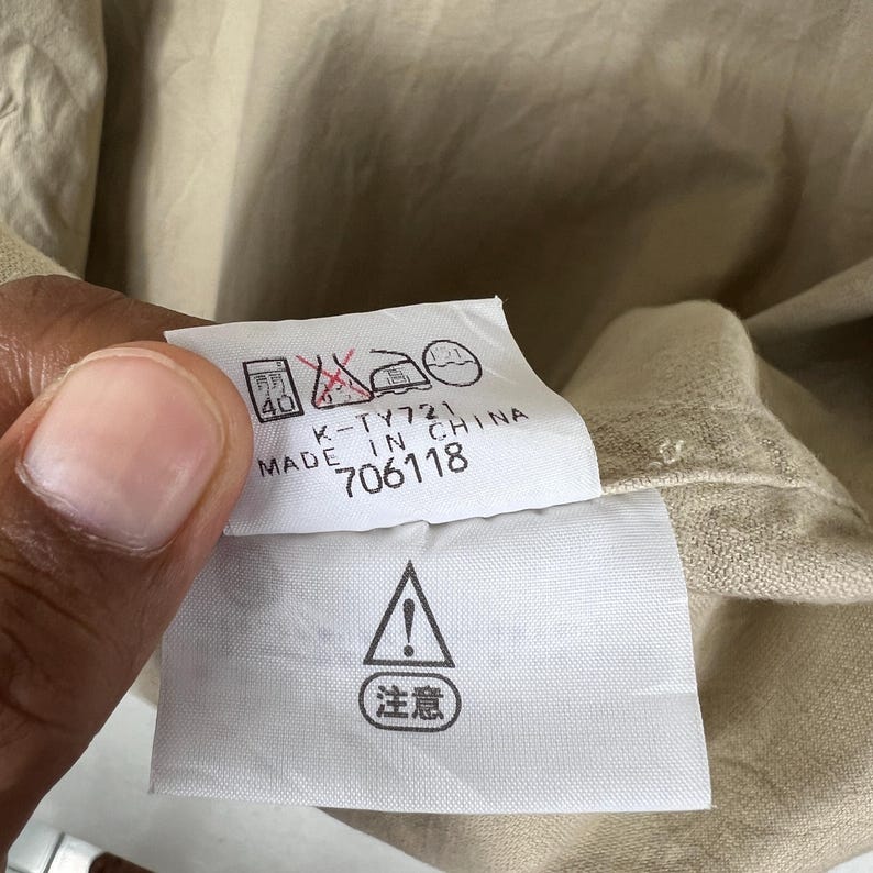 K&ouml;nnte beinhalten: Nahaufnahme eines Kleidungsetiketts mit Pflegesymbolen und Text. Das wei&szlig;e Etikett hat schwarzen Text mit der Aufschrift "K-TY721 MADE IN CHINA 706118". Ein Warnsymbol mit japanischem Text ist ebenfalls zu sehen.