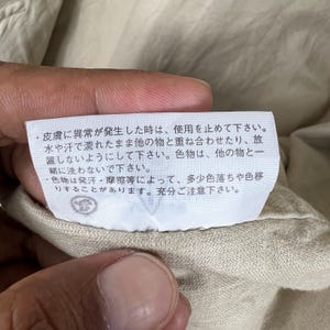 K&ouml;nnte beinhalten: Nahaufnahme eines beigefarbenen Stoffes mit einem wei&szlig;en Etikett. Das Etikett enth&auml;lt japanischen Text, einschlie&szlig;lich Warnhinweisen zu Gebrauch und Pflege. Der Stoff wird von einer Hand gehalten.