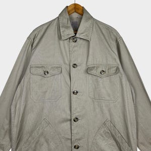 K&ouml;nnte beinhalten: Hellbeige Jacke mit Kragen und Knopfverschluss. Die Jacke hat zwei Brusttaschen und zwei untere Taschen. Das Filson-Markenetikett ist auf der R&uuml;ckseite des Kragens sichtbar. Die Jacke h&auml;ngt an einem Holzb&uuml;gel.