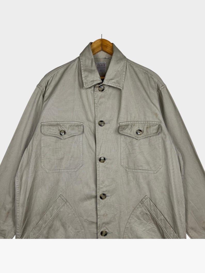 K&ouml;nnte beinhalten: Hellbeige Jacke mit Kragen und Knopfverschluss. Die Jacke hat zwei Brusttaschen und zwei untere Taschen. Das Filson-Markenetikett ist auf der R&uuml;ckseite des Kragens sichtbar. Die Jacke h&auml;ngt an einem Holzb&uuml;gel.