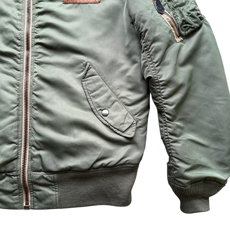 May include: An olive green bomber jacket featuring a central metal zip, a snap-button pocket on the front, and a zipped utility pocket on the sleeve. The garment includes a ribbed knit hem and sleeve cuffs, with a leather patch on the chest.