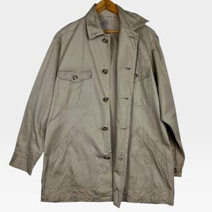 K&ouml;nnte beinhalten: Beige Jacke mit Knopfleiste, zwei Brusttaschen und zwei unteren Taschen. Die Jacke h&auml;ngt an einem Holzb&uuml;gel. Die Jacke hat einen Kragen und lange &Auml;rmel.