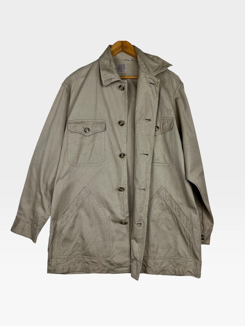 K&ouml;nnte beinhalten: Beige Jacke mit Knopfleiste, zwei Brusttaschen und zwei unteren Taschen. Die Jacke h&auml;ngt an einem Holzb&uuml;gel. Die Jacke hat einen Kragen und lange &Auml;rmel.