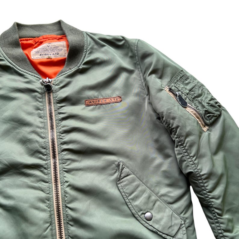 May include: An olive green bomber jacket featuring a bright orange interior lining, a full-length front zip, and a ribbed collar. A brown leather patch with the text AVIREX LTD is on the chest, and a zipped utility pocket is on the sleeve.