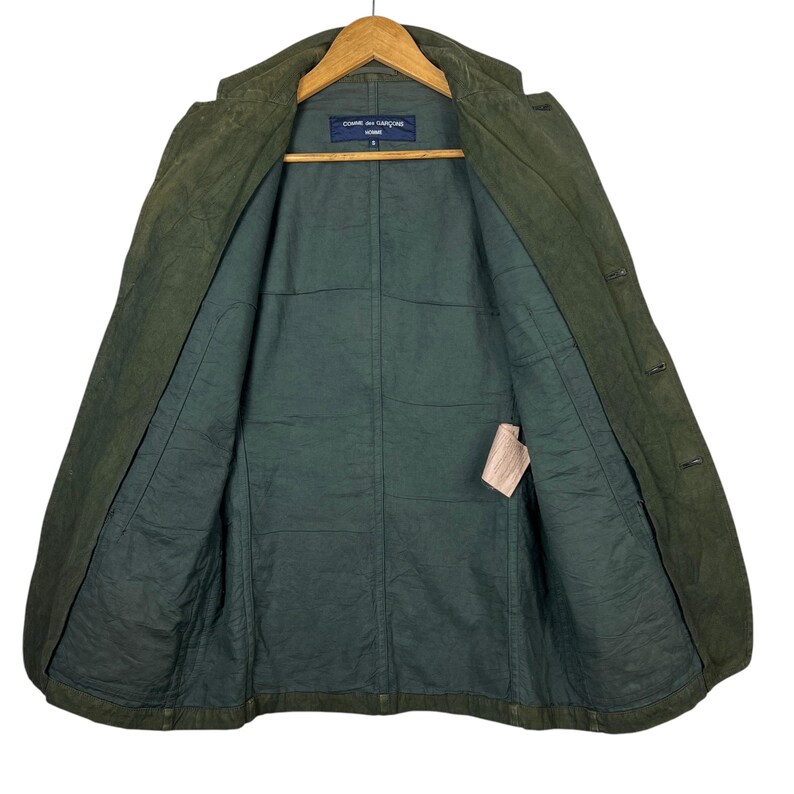 May include: Open olive green jacket with a dark green interior. The jacket is hanging on a wooden hanger. The interior has a label with text. The jacket has buttons and pockets.