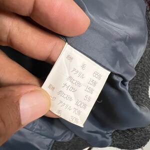 Puede incluir: Primer plano de una etiqueta de ropa con texto japon&eacute;s y porcentajes. La etiqueta blanca con texto negro enumera la composici&oacute;n de la tela: lana, acr&iacute;lico, poli&eacute;ster y nailon. La etiqueta se sostiene contra una tela gris.