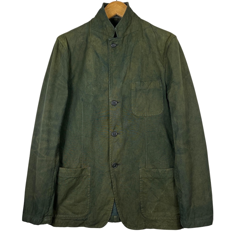 May include: A green jacket with a mandarin collar, three buttons, and two front pockets. The jacket is made of a textured fabric and hangs on a wooden hanger.