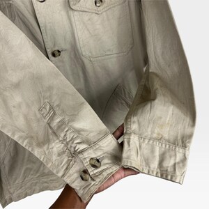 K&ouml;nnte beinhalten: Beigefarbene Jacke mit Knopfverschl&uuml;ssen und zwei Brusttaschen. Die Jacke hat eine gekn&ouml;pfte Manschette am &Auml;rmel. Der Stoff wirkt leicht zerknittert und es gibt Flecken auf dem &Auml;rmel.