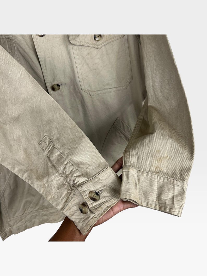 K&ouml;nnte beinhalten: Beigefarbene Jacke mit Knopfverschl&uuml;ssen und zwei Brusttaschen. Die Jacke hat eine gekn&ouml;pfte Manschette am &Auml;rmel. Der Stoff wirkt leicht zerknittert und es gibt Flecken auf dem &Auml;rmel.