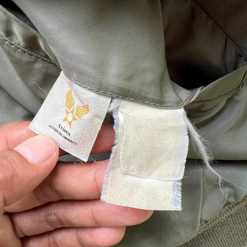 May include: An olive green jacket interior featuring a white fabric tag with the gold Avirex logo and the text AUTHENTIC PRODUCT printed in black. A second blank, frayed white fabric tag is attached next to the branded label on the lining.