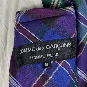 Puede incluir: Una chaqueta de cuadros azules, morados y blancos con una etiqueta negra que dice "COMME des GARCONS* HOMME PLUS M".