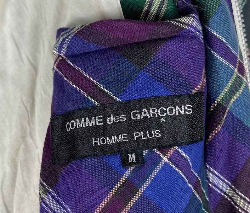 Puede incluir: Una chaqueta de cuadros azules, morados y blancos con una etiqueta negra que dice "COMME des GARCONS* HOMME PLUS M".