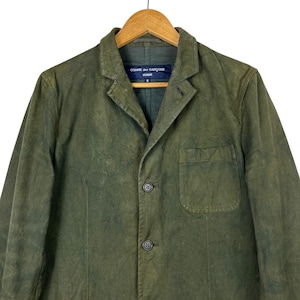 May include: Olive green blazer with a notched lapel, three buttons, and a chest pocket. The jacket is hanging on a wooden hanger. The label reads "COMME des GAR&Ccedil;ONS HOMME".