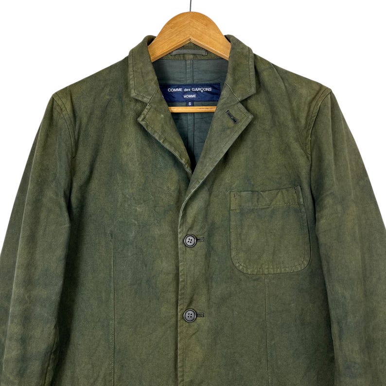 May include: Olive green blazer with a notched lapel, three buttons, and a chest pocket. The jacket is hanging on a wooden hanger. The label reads "COMME des GAR&Ccedil;ONS HOMME".