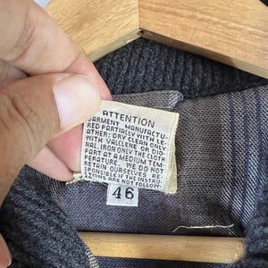 Puede incluir: Primer plano de una prenda gris oscuro con una etiqueta. La etiqueta dice "ATTENTION" y proporciona instrucciones de cuidado, incluyendo limpieza en seco y planchado. El n&uacute;mero "46" est&aacute; impreso.