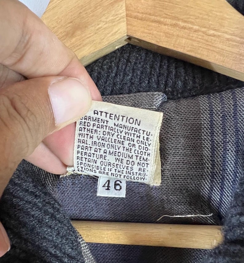 Puede incluir: Primer plano de una prenda gris oscuro con una etiqueta. La etiqueta dice "ATTENTION" y proporciona instrucciones de cuidado, incluyendo limpieza en seco y planchado. El n&uacute;mero "46" est&aacute; impreso.