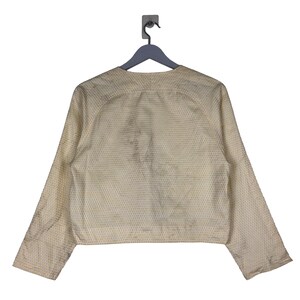 K&ouml;nnte beinhalten: Eine cremefarbene Jacke mit einem dezenten wei&szlig;en und cremefarbenen Muster. Die Jacke hat lange &Auml;rmel und einen Knopfverschluss vorne.