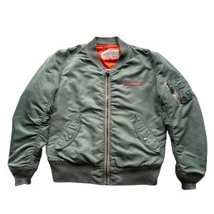May include: An olive green bomber jacket featuring a bright orange interior lining, a full-length silver metal front zip, ribbed cuffs and hem, two front snap-button pockets, and a zipped utility pocket on the left sleeve with a leather pull.
