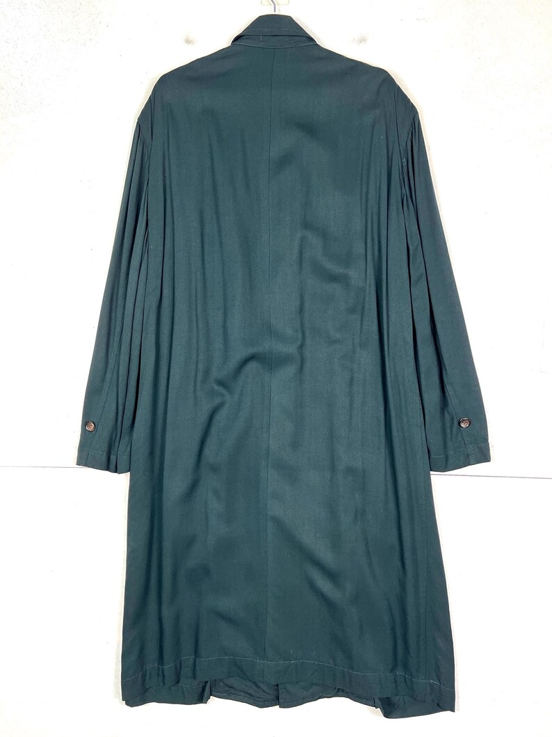 Może przedstawiać: Długi, ciemnozielony płaszcz z zapięciem na guziki i długimi rękawami. Płaszcz ma klasyczny kr&oacute;j z kołnierzem i dopasowanym krojem. Tkanina wydaje się być materiałem o średniej gramaturze, odpowiednim na chłodniejszą pogodę.