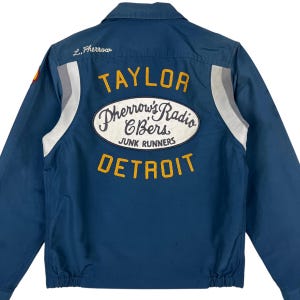 May include: A blue work jacket featuring a large oval patch on the back with the text Pherrow's Radio CBers Junk Runners. Above the patch is the name Taylor in yellow embroidery, and below is Detroit. The name L. Pherrow is embroidered in white.