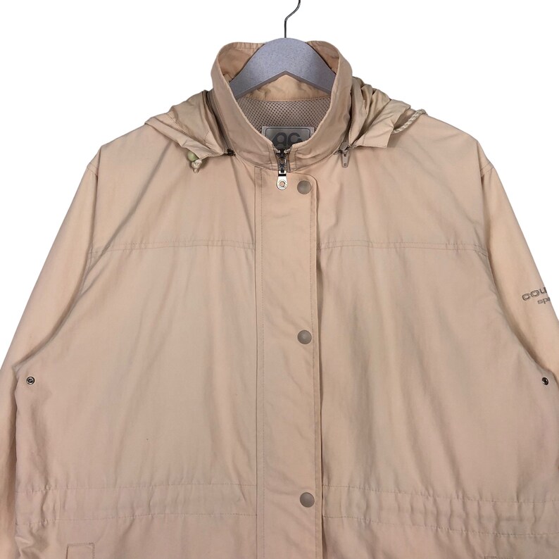 May include: A beige hooded jacket with a zipper closure and button accents. The jacket has a drawstring waist and a logo on the right sleeve that reads "Columbia Sportswear".