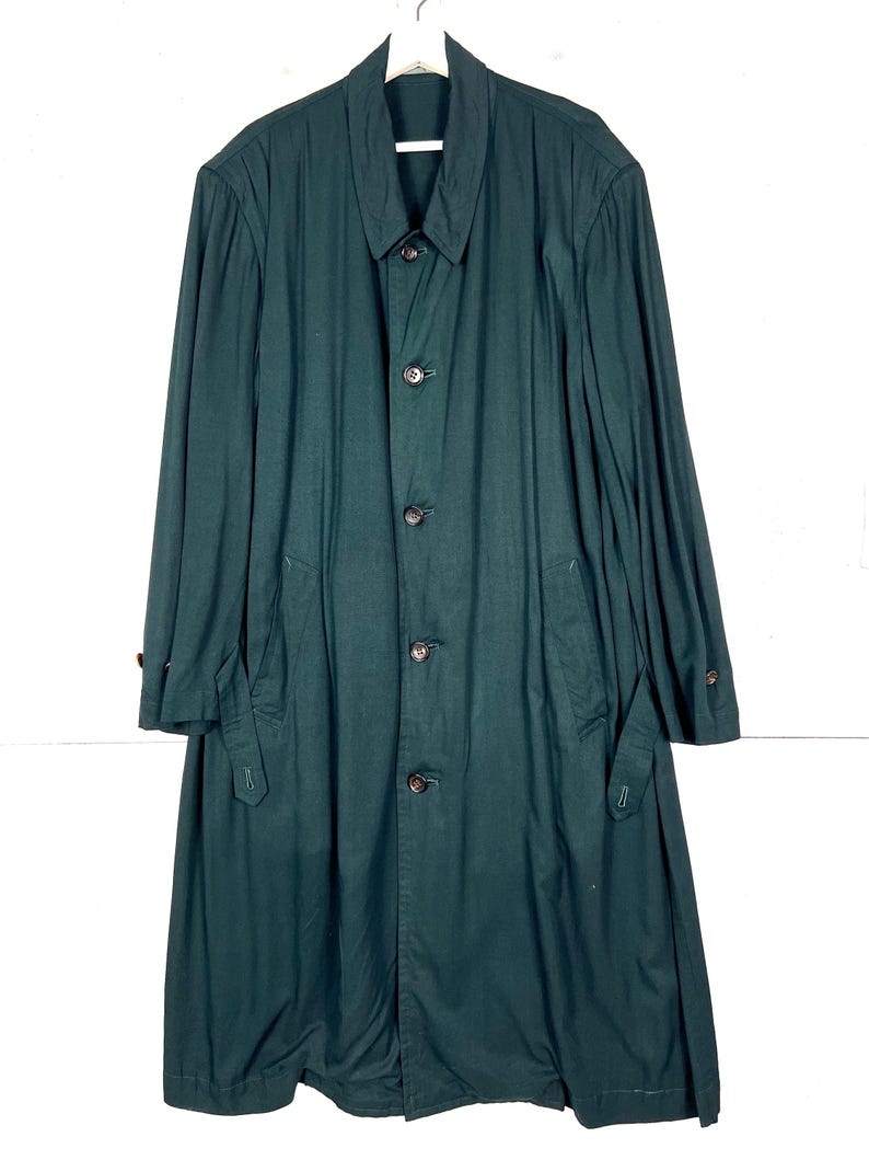 Może przedstawiać: Długi, ciemnozielony płaszcz z zapięciem na guziki, kołnierzem i kieszeniami. Płaszcz wykonany jest z jednolitego materiału. Płaszcz wisi na białym wieszaku.