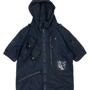 Puede incluir: Una chaqueta negra de manga corta con capucha y múltiples bolsillos y cremalleras. La chaqueta tiene un parche de plástico transparente con la palabra "Puma" en él.