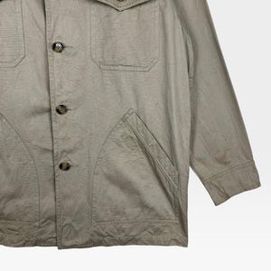 K&ouml;nnte beinhalten: Beige Jacke mit vier Fronttaschen und Knopfverschl&uuml;ssen. Die Jacke hat einen Kragen und lange &Auml;rmel. Der Stoff scheint eine leichte Baumwoll- oder Leinenmischung mit leicht strukturierter Optik zu sein.