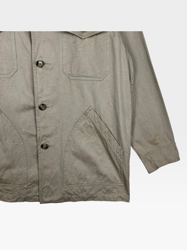 K&ouml;nnte beinhalten: Beige Jacke mit vier Fronttaschen und Knopfverschl&uuml;ssen. Die Jacke hat einen Kragen und lange &Auml;rmel. Der Stoff scheint eine leichte Baumwoll- oder Leinenmischung mit leicht strukturierter Optik zu sein.