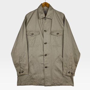 K&ouml;nnte beinhalten: Beigefarbene Knopfjacke mit zwei Brusttaschen und zwei unteren Taschen. Die Jacke hat lange &Auml;rmel mit gekn&ouml;pften Manschetten und einen Kragen. Sie h&auml;ngt an einem Holzb&uuml;gel.