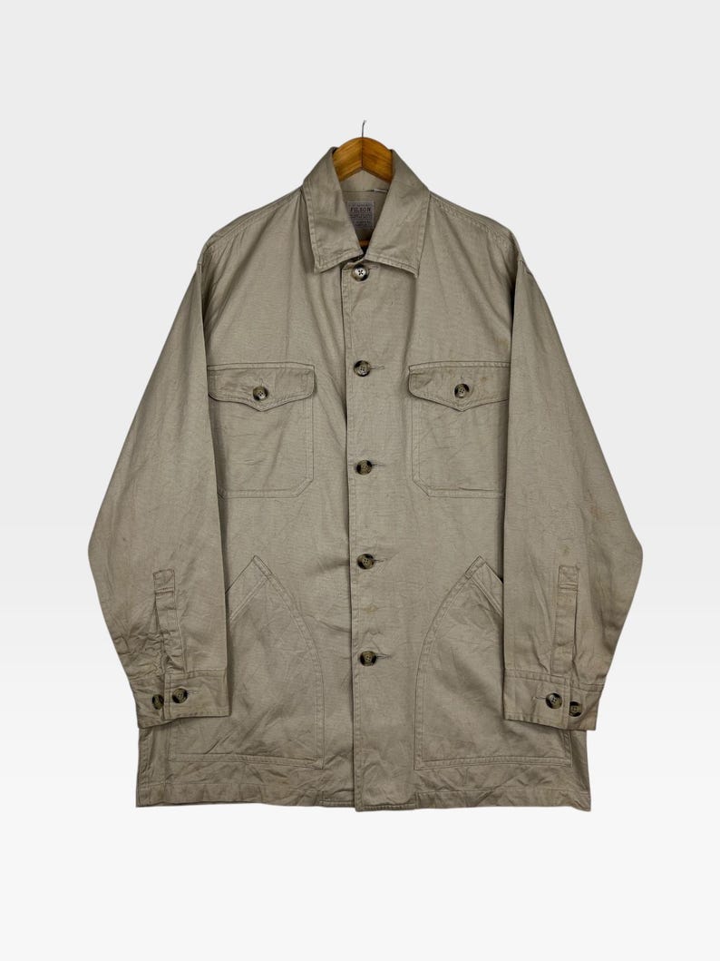 K&ouml;nnte beinhalten: Beigefarbene Knopfjacke mit zwei Brusttaschen und zwei unteren Taschen. Die Jacke hat lange &Auml;rmel mit gekn&ouml;pften Manschetten und einen Kragen. Sie h&auml;ngt an einem Holzb&uuml;gel.