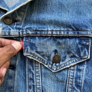 May include: Close-up of a blue denim jacket with a pocket and metal buttons. The jacket has a red tag with white lettering. The denim is faded and worn, with visible stitching and a classic design.
