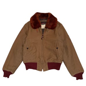 May include: A brown bomber jacket with a faux fur collar and a burgundy ribbed hem and cuffs. The jacket has a zipper closure and two front pockets. The jacket has a label on the inside of the collar.