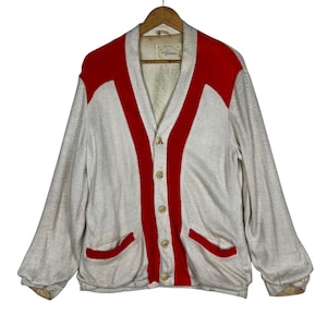 May include: A vintage light grey cardigan with red detailing. The sweater has a V-neck, button closure, and two front pockets. Red accents are on the shoulders and down the front. The label reads "St. Andrews".