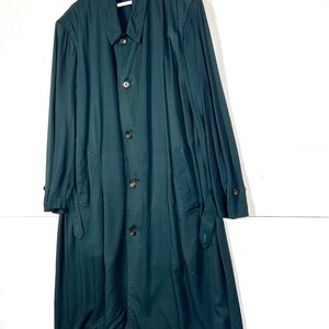 Może przedstawiać: Długi, ciemnozielony płaszcz z zapięciem na guziki i kołnierzem, wiszący na białym wieszaku. Płaszcz ma długie rękawy i kieszenie. Płaszcz jest wykonany z gładkiej tkaniny.