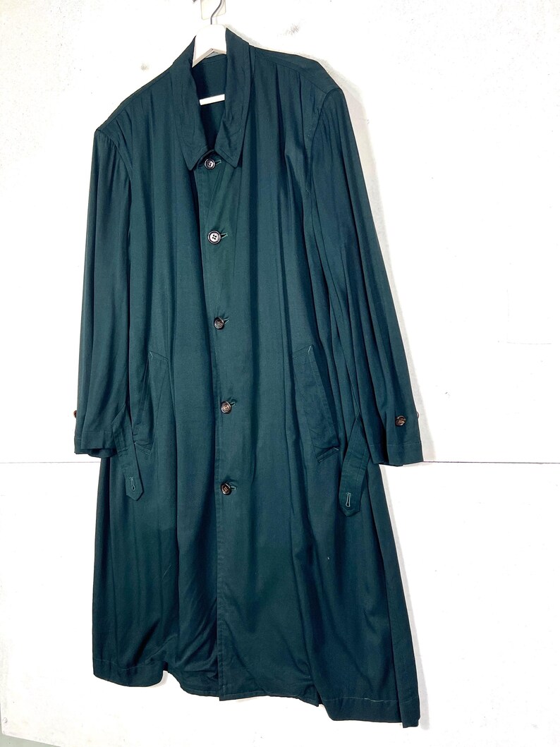 Może przedstawiać: Długi, ciemnozielony płaszcz z zapięciem na guziki i kołnierzem, wiszący na białym wieszaku. Płaszcz ma długie rękawy i kieszenie. Płaszcz jest wykonany z gładkiej tkaniny.