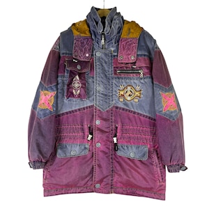 May include: A multi-coloured jacket featuring a purple and grey colour-blocked design with a hood. The jacket includes multiple pockets, a front zip closure with a B-shaped pull, and embroidered patches on the chest and sleeves in pink and gold.
