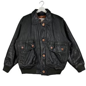 May include: Black leather bomber jacket with a button-up front, two chest pockets, and two lower pockets. The jacket has a collared neckline, ribbed cuffs, and a ribbed hem. The buttons and pocket details are a light brown color.