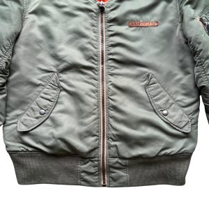 May include: An olive green bomber jacket featuring a central metal zip, two slanted front pockets with snap button closures, a ribbed hem, and a rectangular brown leather patch on the chest with the embossed text AVIREX LTD.