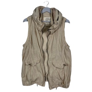 May include: A beige sleeveless vest with a zipper closure and a high collar. The vest has two pockets on the front and a drawstring at the bottom.