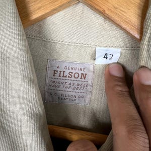 K&ouml;nnte beinhalten: Nahaufnahme eines beigefarbenen Filson-Hemdetiketts mit dem Text "A GENUINE FILSON" und "MIGHT AS WELL HAVE THE BEST". Das Etikett enth&auml;lt auch "C.C. FILSON CO. SEATTLE". Ein Gr&ouml;&szlig;enetikett 42 ist sichtbar.