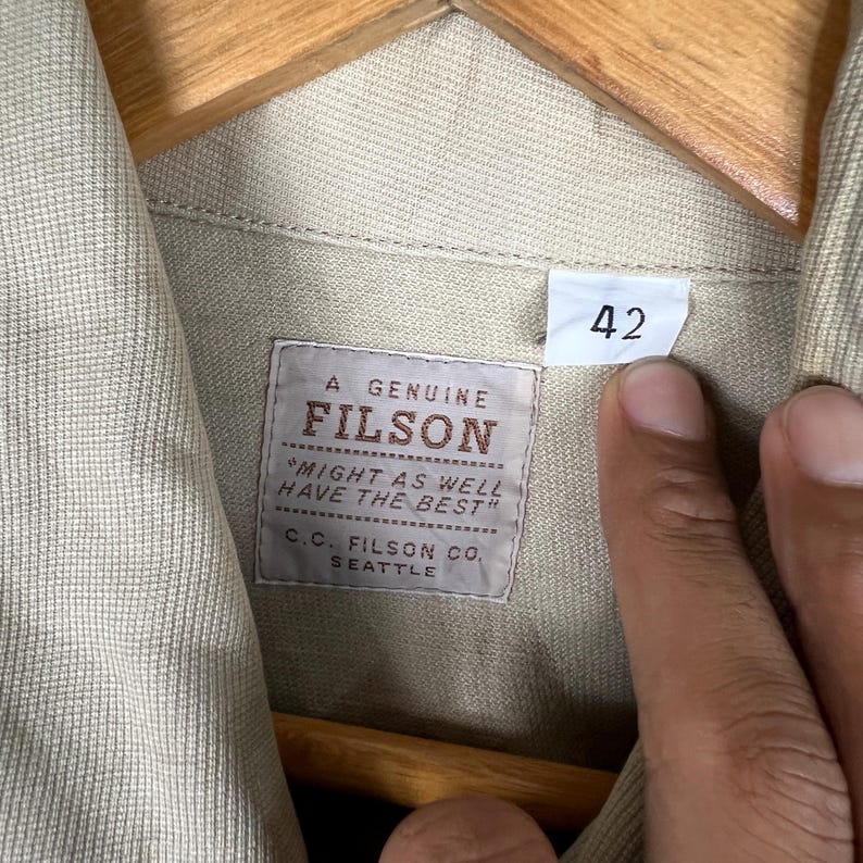 K&ouml;nnte beinhalten: Nahaufnahme eines beigefarbenen Filson-Hemdetiketts mit dem Text "A GENUINE FILSON" und "MIGHT AS WELL HAVE THE BEST". Das Etikett enth&auml;lt auch "C.C. FILSON CO. SEATTLE". Ein Gr&ouml;&szlig;enetikett 42 ist sichtbar.