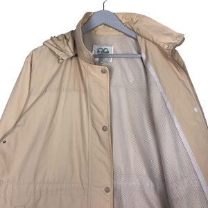 May include: A beige hooded jacket with a zipper and button closure. The jacket has a mesh lining and a small logo on the left chest.