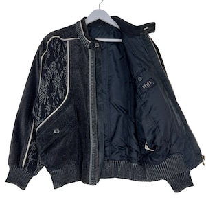 May include: A dark gray corduroy bomber jacket with a unique design. The jacket features a textured pattern on the left shoulder and a contrasting white trim. It has a zip closure, two pockets, and the brand name "RAIKA" visible on the inside.