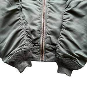 May include: An olive green bomber jacket featuring a central metal zip closure, long sleeves with gathered fabric detailing, and ribbed knit cuffs at the wrists. The jacket is constructed from a smooth, slightly reflective synthetic material.