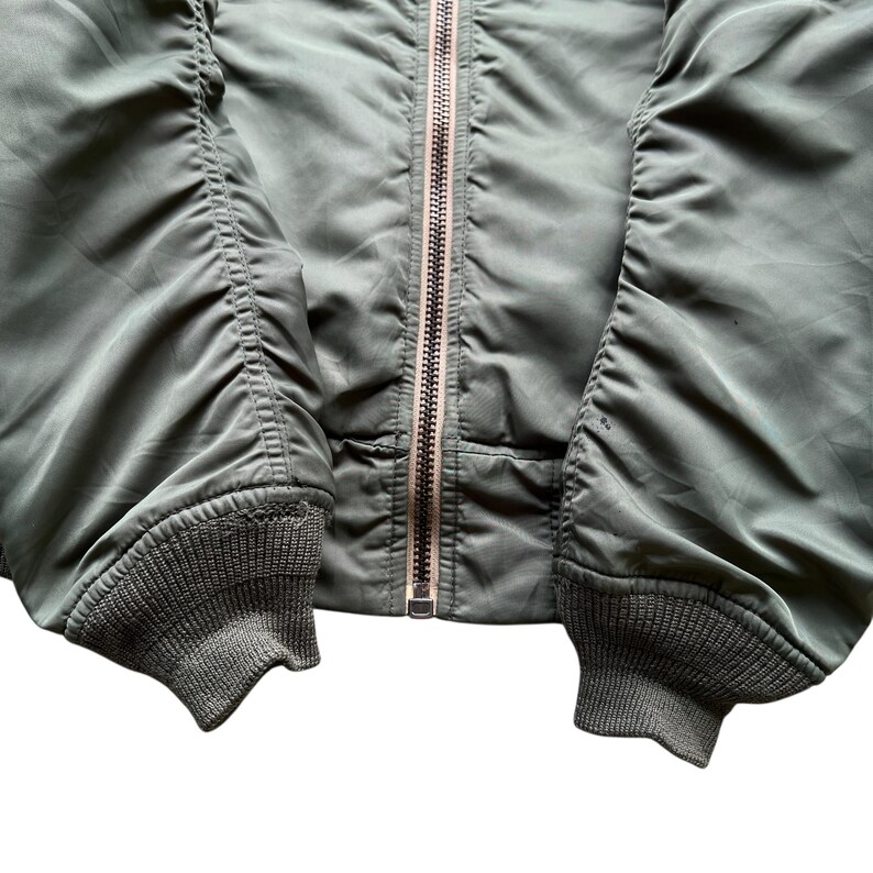 May include: An olive green bomber jacket featuring a central metal zip closure, long sleeves with gathered fabric detailing, and ribbed knit cuffs at the wrists. The jacket is constructed from a smooth, slightly reflective synthetic material.