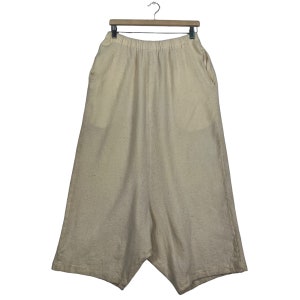 Puede incluir: Pantalones color crema de pierna ancha con cintura elástica y bolsillos. Los pantalones están hechos de una tela texturizada y cuelgan de una percha de madera. El diseño presenta un dobladillo asimétrico único.