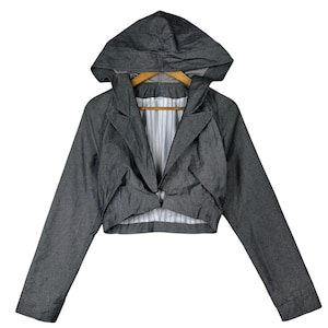 Pode incluir: Um blazer cinza escuro cortado com capuz. O blazer tem mangas compridas e lapelas entalhadas. O forro interior é branco com listras verticais. O casaco está pendurado em um cabide de madeira.
